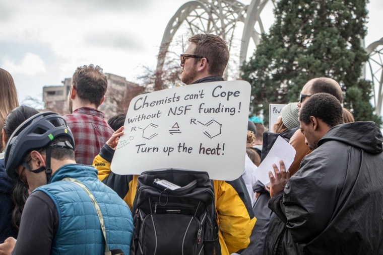 In a crowd of people, a person holds a sign that says "Chemists can't cope without NSF funding, turn up the heat!"
