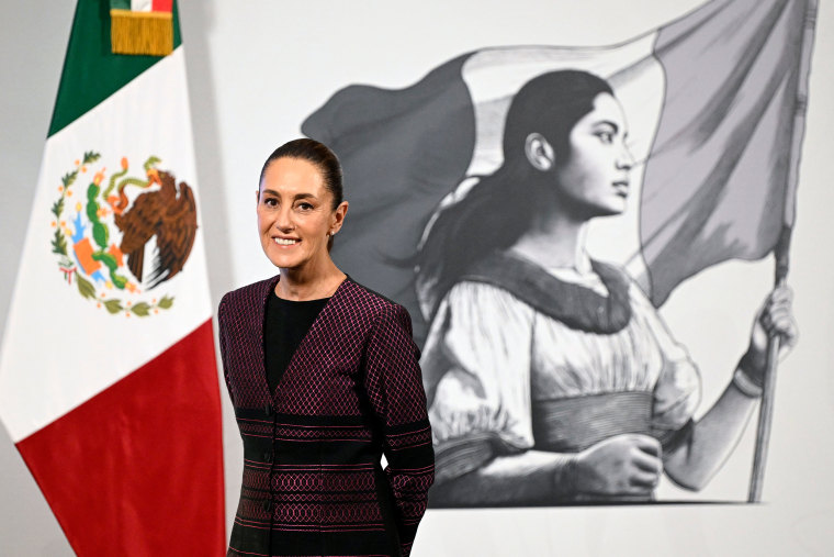 Claudia Sheinbaum smiling in front a Mexican flag and mural