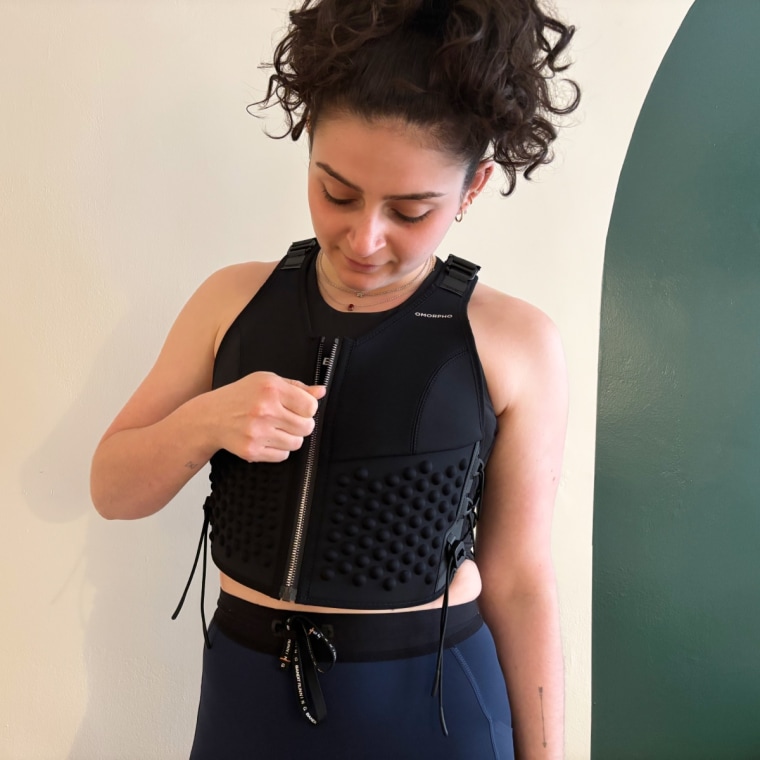 A woman wearing and zipping up Omorpho’s black G-Vest while standing in front of a white and green wall.